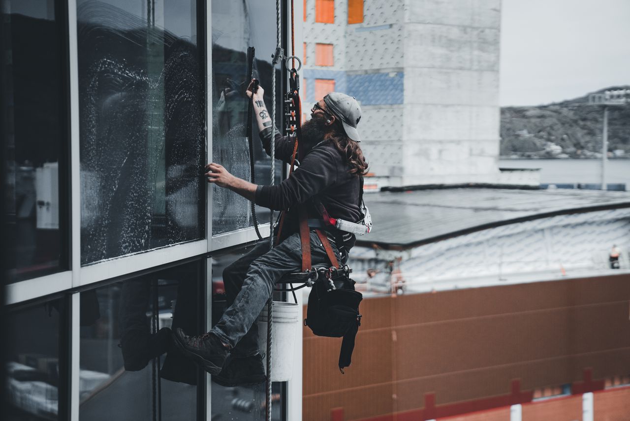 gallery-1 A man cleaning skyscraper windows with a harness, showcasing urban high-rise work.